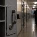Close-up of a secured prison cell door and dim corridor, with a blurred correctional officer walking in the background.