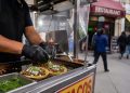 Close-up of a sidewalk food cart preparing tacos, with blurred pedestrians and a restaurant storefront in the background.