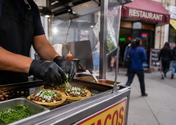 Close-up of a sidewalk food cart preparing tacos, with blurred pedestrians and a restaurant storefront in the background.