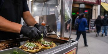 Close-up of a sidewalk food cart preparing tacos, with blurred pedestrians and a restaurant storefront in the background.
