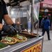 Close-up of a sidewalk food cart preparing tacos, with blurred pedestrians and a restaurant storefront in the background.