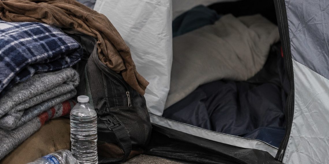 Close-up of a sidewalk tent entrance beside folded blankets, bottled water and personal belongings.