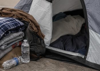 Close-up of a sidewalk tent entrance beside folded blankets, bottled water and personal belongings.
