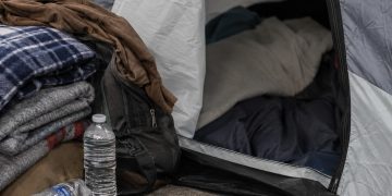 Close-up of a sidewalk tent entrance beside folded blankets, bottled water and personal belongings.