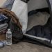 Close-up of a sidewalk tent entrance beside folded blankets, bottled water and personal belongings.