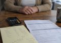 Close-up of a smartphone beside handwritten account notes and a bank statement, with a blurred older adult seated across the table.