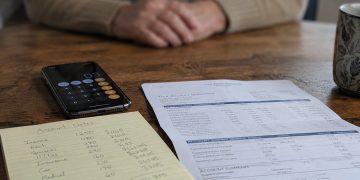 Close-up of a smartphone beside handwritten account notes and a bank statement, with a blurred older adult seated across the table.