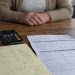 Close-up of a smartphone beside handwritten account notes and a bank statement, with a blurred older adult seated across the table.