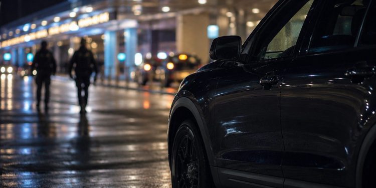 Close-up of airport arrival-area lights and an unmarked law-enforcement vehicle at night, with two blurred officers walking in the background.