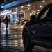 Close-up of airport arrival-area lights and an unmarked law-enforcement vehicle at night, with two blurred officers walking in the background.