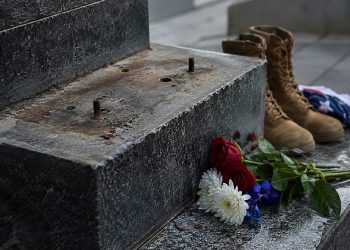 Close-up of an empty bronze statue base at a military memorial with flowers and boots nearby.
