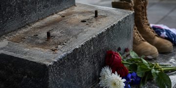Close-up of an empty bronze statue base at a military memorial with flowers and boots nearby.