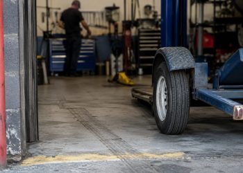 Close-up of an empty vehicle repair bay with tire marks and a tow dolly wheel near the entrance, with a blurred mechanic standing farther inside.