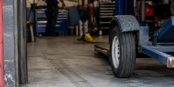 Close-up of an empty vehicle repair bay with tire marks and a tow dolly wheel near the entrance, with a blurred mechanic standing farther inside.