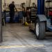 Close-up of an empty vehicle repair bay with tire marks and a tow dolly wheel near the entrance, with a blurred mechanic standing farther inside.