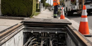 Close-up of an open underground utility vault with electrical cables and warning cones, with a blurred utility worker standing nearby.