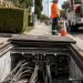 Close-up of an open underground utility vault with electrical cables and warning cones, with a blurred utility worker standing nearby.