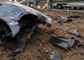 Close-up of bent vehicle metal and broken plastic debris resting on dirt beside a roadway.