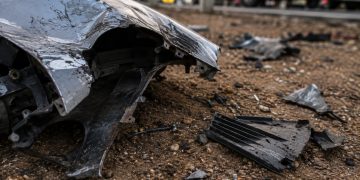 Close-up of bent vehicle metal and broken plastic debris resting on dirt beside a roadway.