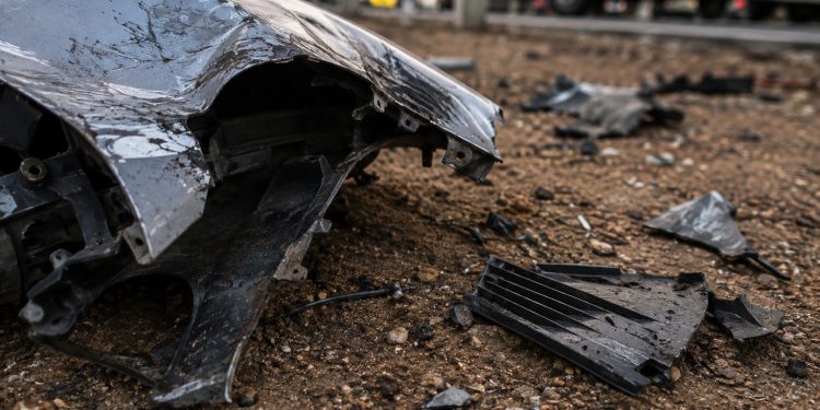 Close-up of bent vehicle metal and broken plastic debris resting on dirt beside a roadway.
