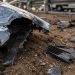 Close-up of bent vehicle metal and broken plastic debris resting on dirt beside a roadway.