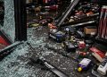 Close-up of broken storefront glass, scattered auto parts and damaged shelving on a dark floor.