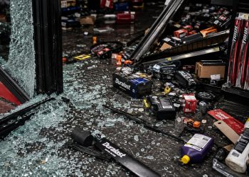Close-up of broken storefront glass, scattered auto parts and damaged shelving on a dark floor.