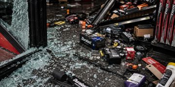Close-up of broken storefront glass, scattered auto parts and damaged shelving on a dark floor.