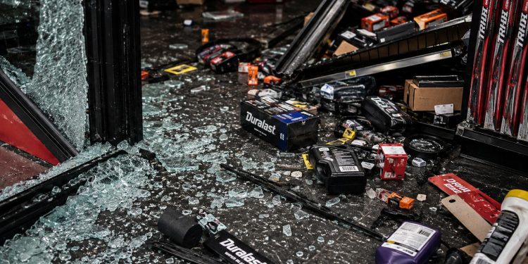 Close-up of broken storefront glass, scattered auto parts and damaged shelving on a dark floor.