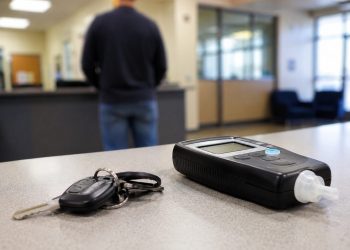 Close-up of car keys beside a sobriety testing device on a CHP office counter, with a blurred person standing in the lobby background.