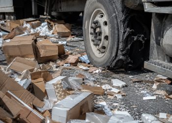 Close-up of cardboard boxes and broken cargo scattered across freeway pavement beside a damaged truck tire.