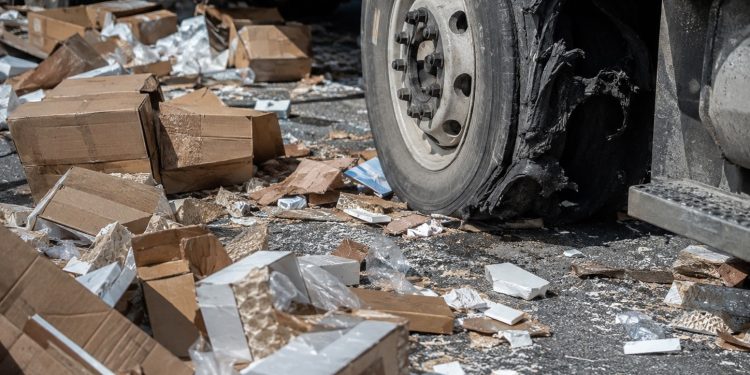 Close-up of cardboard boxes and broken cargo scattered across freeway pavement beside a damaged truck tire.
