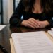 Close-up of court documents and a pen on a table, with a blurred woman seated in the background.
