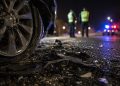 Close-up of crash debris beside a damaged vehicle wheel at night, with two blurred investigators and distant emergency lights in the background.