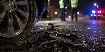 Close-up of crash debris beside a damaged vehicle wheel at night, with two blurred investigators and distant emergency lights in the background.