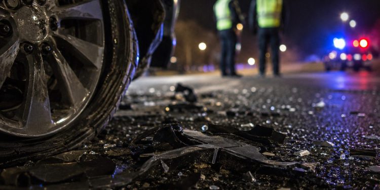 Close-up of crash debris beside a damaged vehicle wheel at night, with two blurred investigators and distant emergency lights in the background.