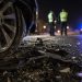 Close-up of crash debris beside a damaged vehicle wheel at night, with two blurred investigators and distant emergency lights in the background.
