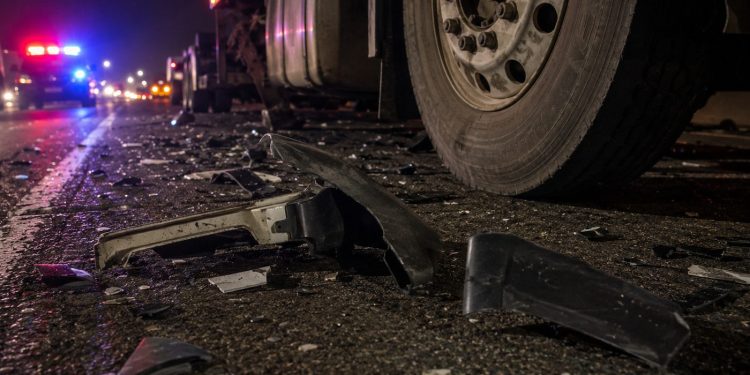 Close-up of damaged vehicle debris beside a big rig tire on freeway pavement at night.