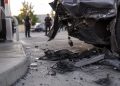 Close-up of damaged vehicle panels and scattered debris near a gas station driveway, with blurred officers standing farther back.