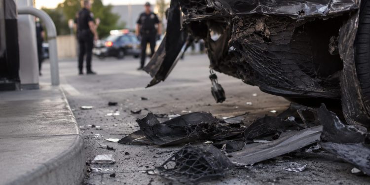 Close-up of damaged vehicle panels and scattered debris near a gas station driveway, with blurred officers standing farther back.