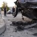 Close-up of damaged vehicle panels and scattered debris near a gas station driveway, with blurred officers standing farther back.