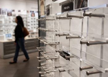 Close-up of empty designer eyeglass display stands inside an optical store, with a blurred shopper walking past in the background.