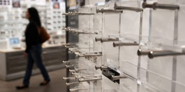 Close-up of empty designer eyeglass display stands inside an optical store, with a blurred shopper walking past in the background.