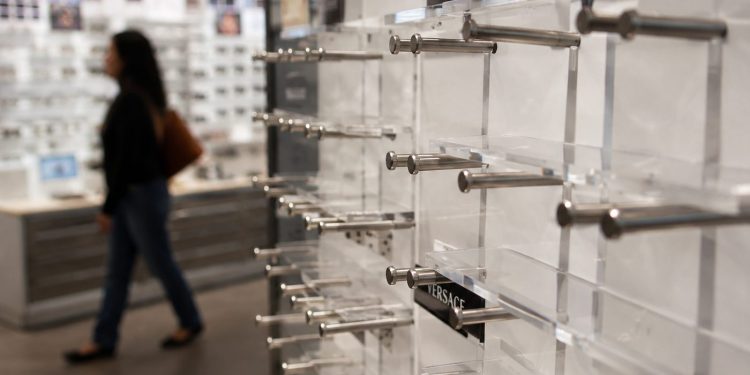 Close-up of empty designer eyeglass display stands inside an optical store, with a blurred shopper walking past in the background.