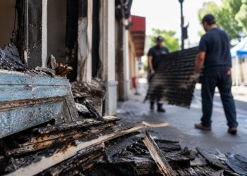 Close-up of fire-damaged storefront materials being cleared from a sidewalk, with two blurred workers carrying debris in the background.