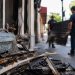 Close-up of fire-damaged storefront materials being cleared from a sidewalk, with two blurred workers carrying debris in the background.