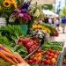 Close-up of fresh produce and flowers arranged beneath farmers market tents along a pedestrian shopping street.