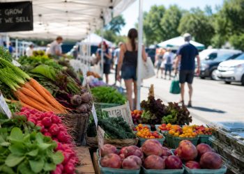 Close-up of fresh produce beneath farmers market tents beside a parking area, with a few blurred shoppers walking between stalls.