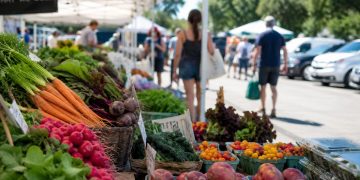 Close-up of fresh produce beneath farmers market tents beside a parking area, with a few blurred shoppers walking between stalls.