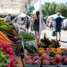 Close-up of fresh produce beneath farmers market tents beside a parking area, with a few blurred shoppers walking between stalls.